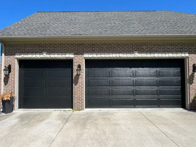 Two black garage doors on a brick house with a concrete driveway and blue sky.