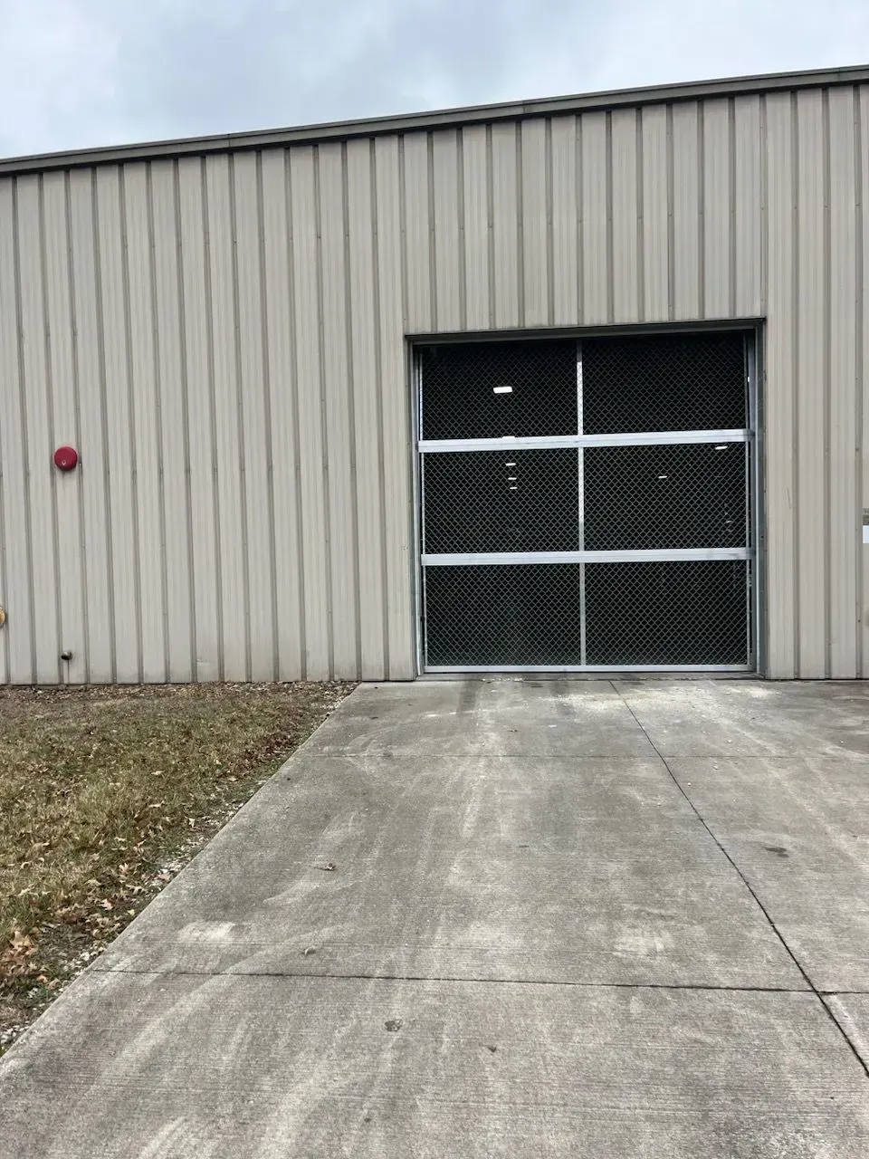Gray industrial building with a metal garage door and concrete driveway, beside a patch of grass