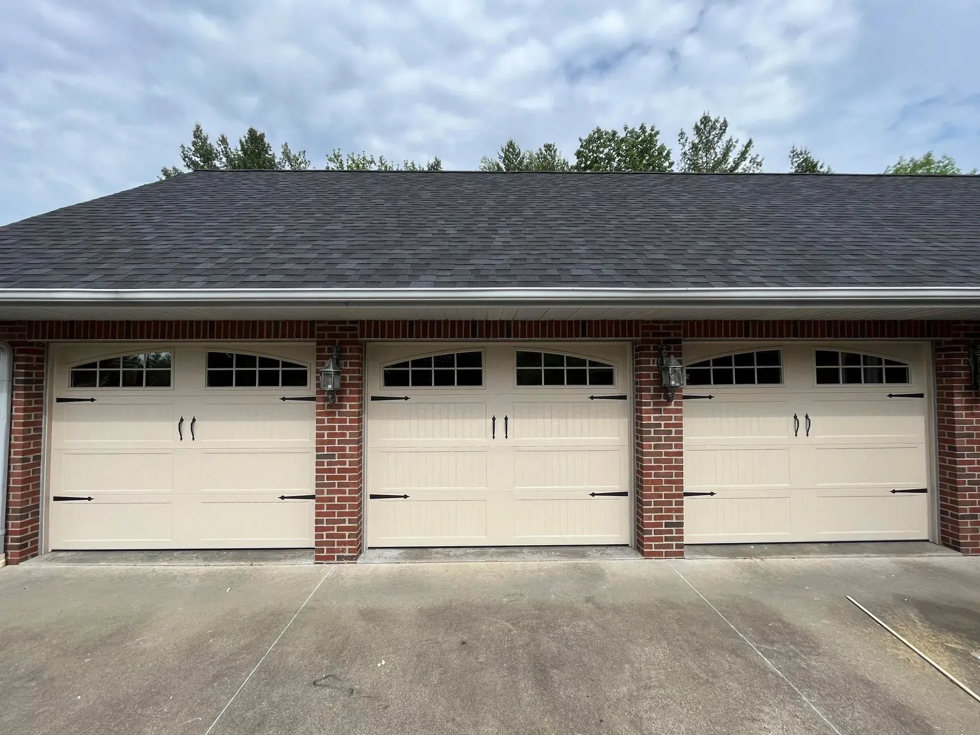 Three beige garage doors in a brick building with a dark shingled roof and concrete driveway