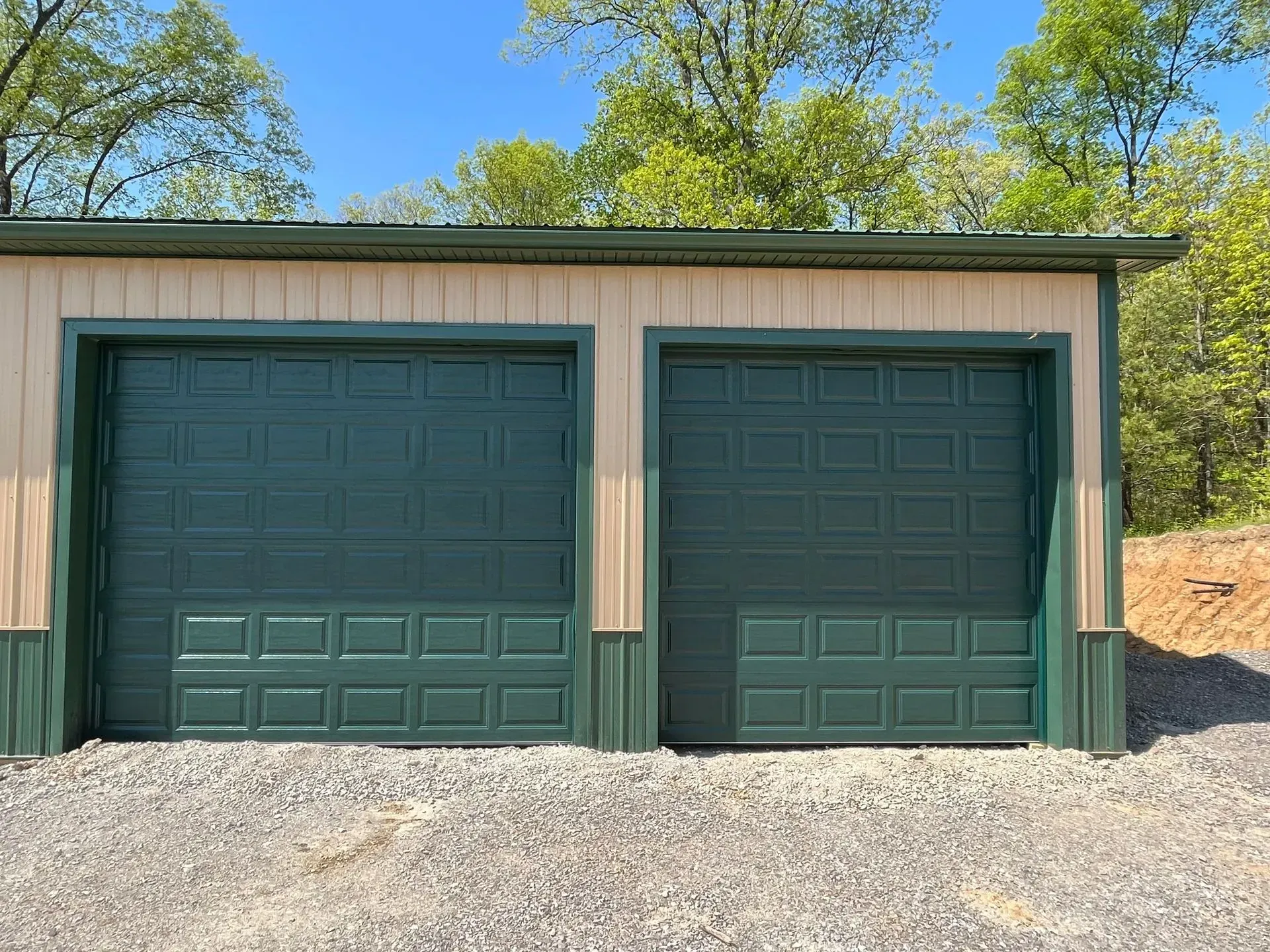 Two dark green garage doors on a beige detached garage with a gravel driveway and trees behind it