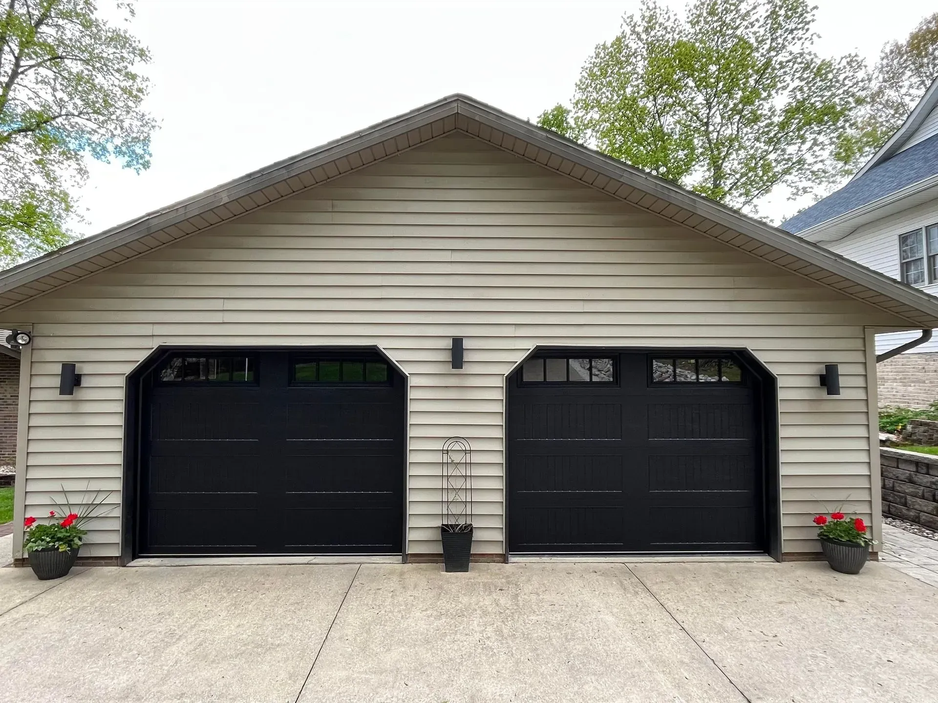 Two-car garage with beige siding, black doors, and flower pots by the driveway