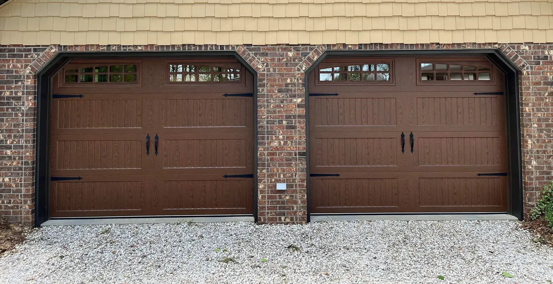 Two brown paneled garage doors set in a brick wall with small windows above and gravel in front