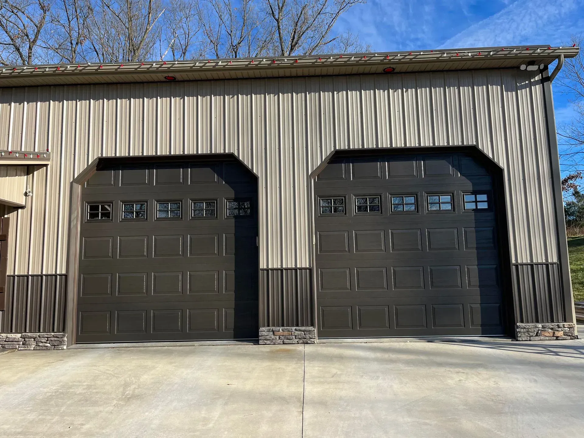 Two black garage doors on a beige metal building with a concrete driveway and blue sky.