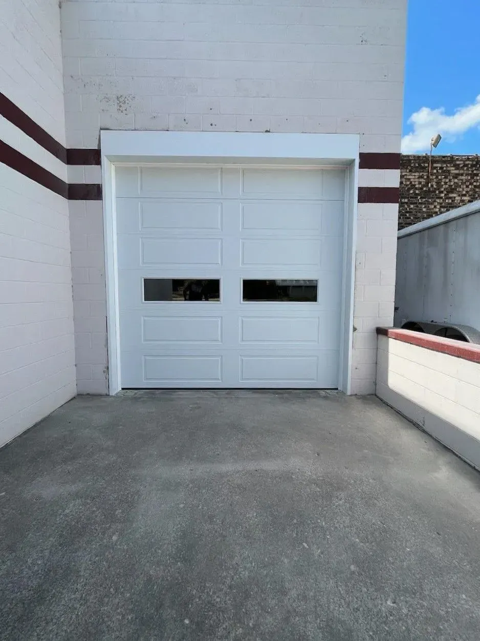 White garage door with two small windows in a narrow concrete driveway beside a building.