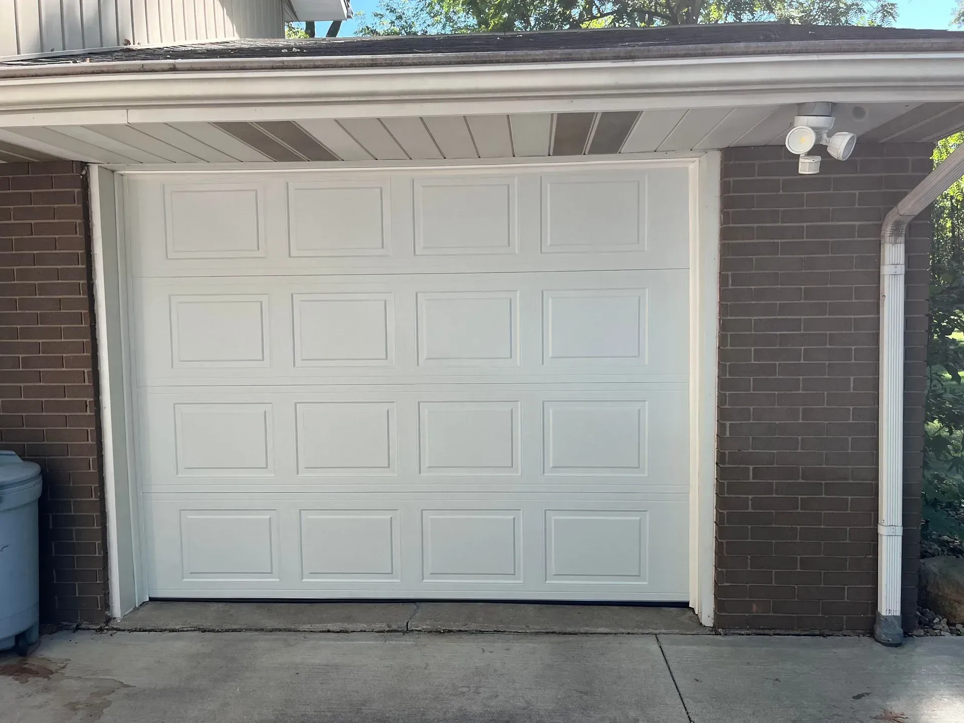 White garage door under a carport attached to a brick house, with a trash bin on the left.