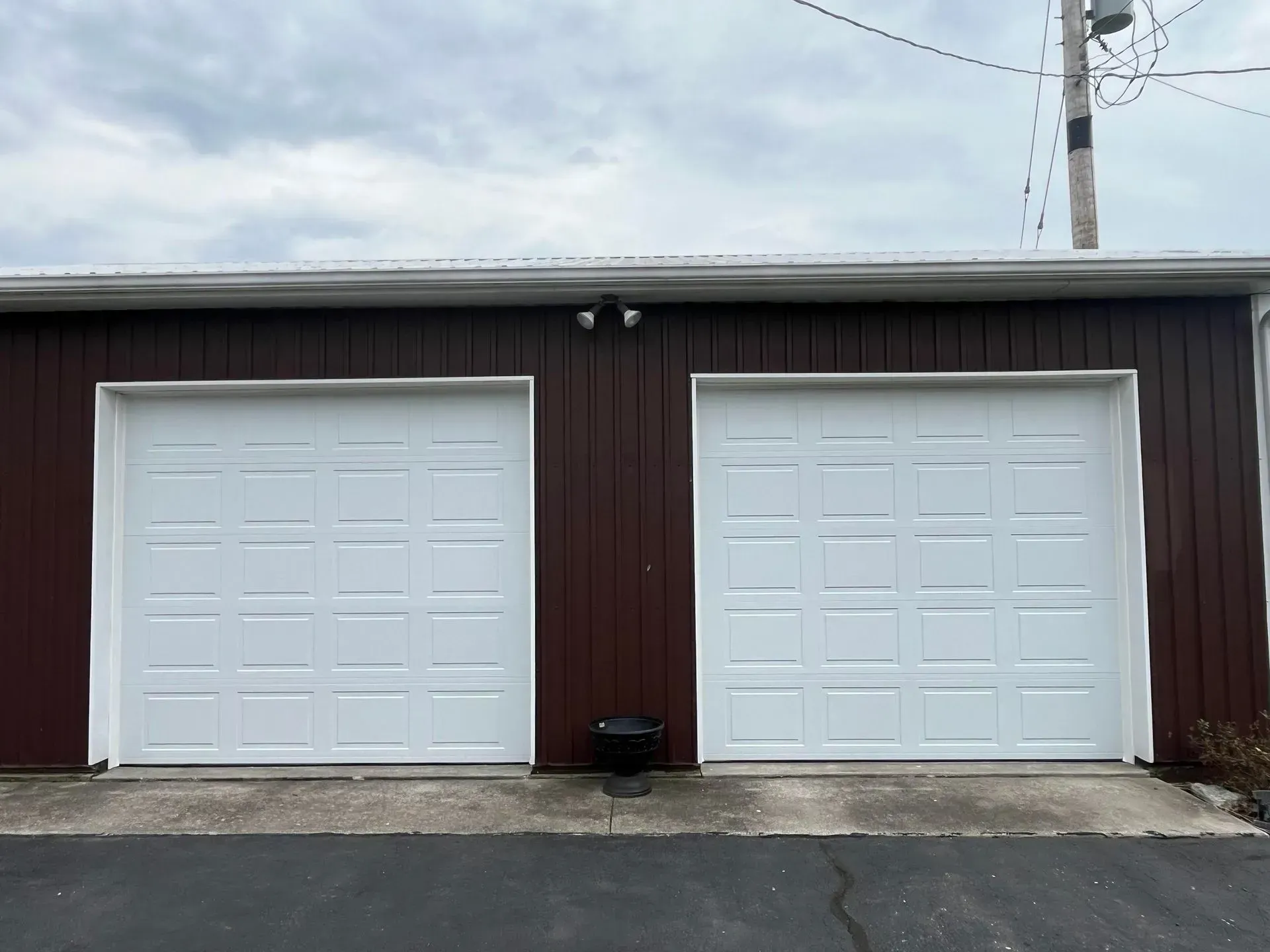 Two white garage doors on a dark brown building with a concrete driveway and utility pole overhead