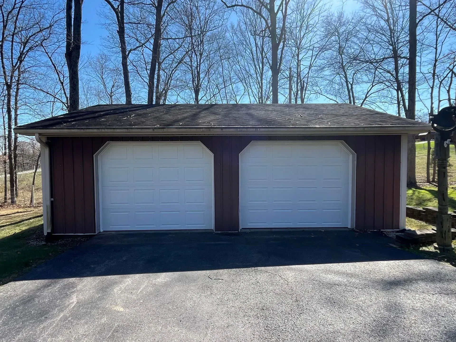 Two-car garage with white doors and brown siding in a wooded driveway.