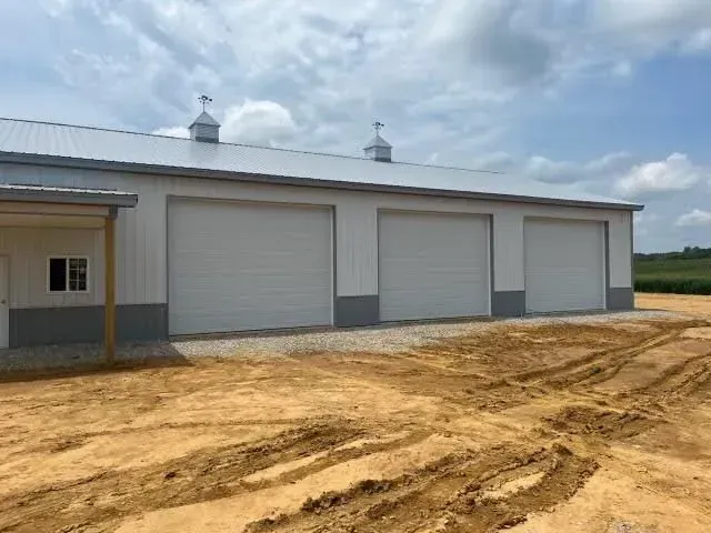 Large white metal garage building with three roll-up doors on a dirt lot under a cloudy sky