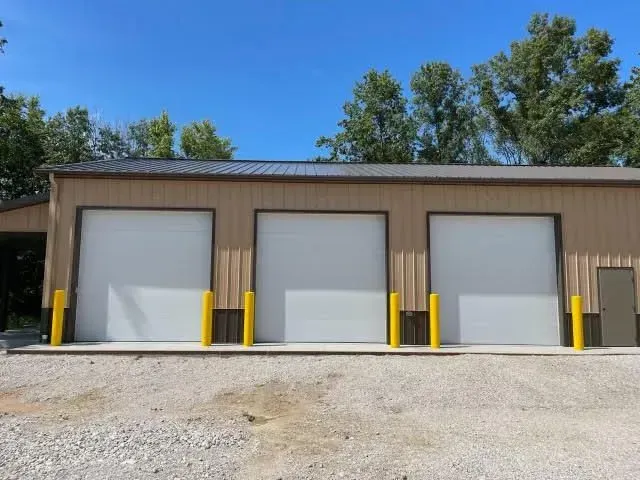 Three white garage doors on a tan industrial building with yellow bollards and trees behind it