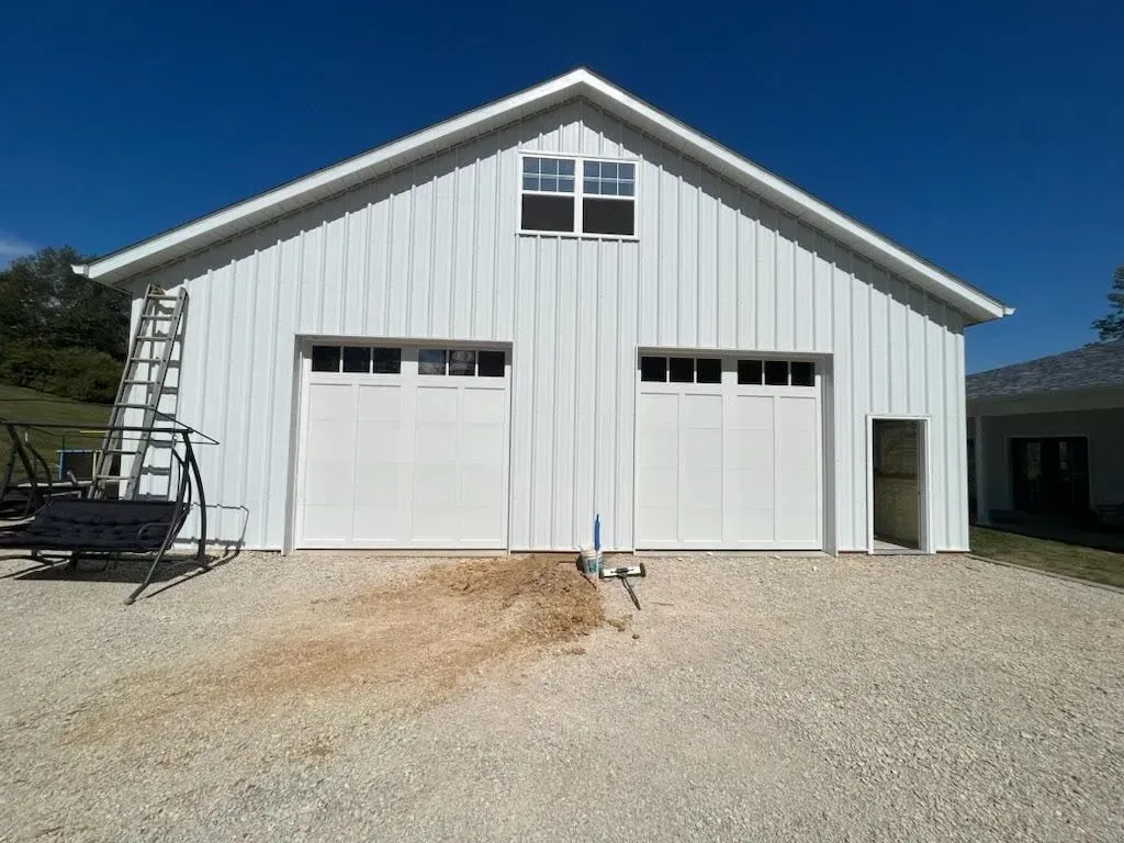 White metal barn with two garage doors and a central loft window on a gravel lot under a clear blue sky