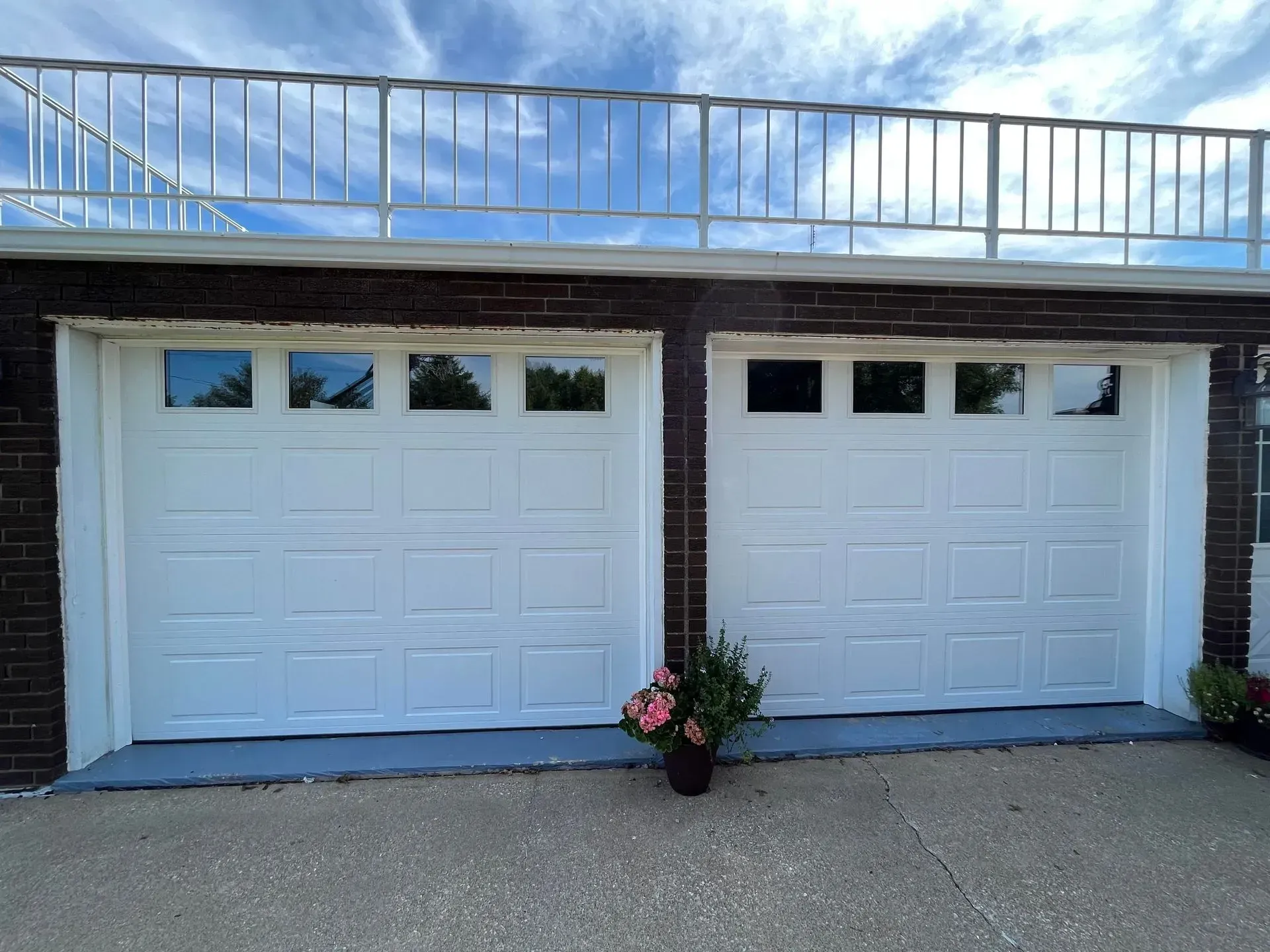 Two white garage doors on a brick house with a railing balcony above and flower pots in front