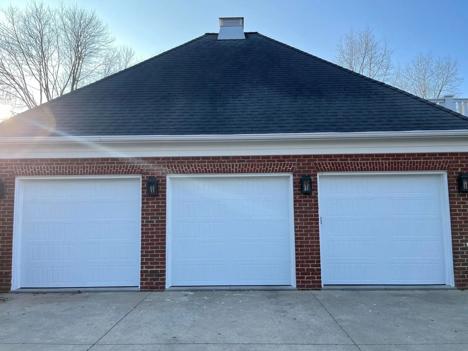 Three white garage doors on a red brick building with a dark roof and driveway in front.