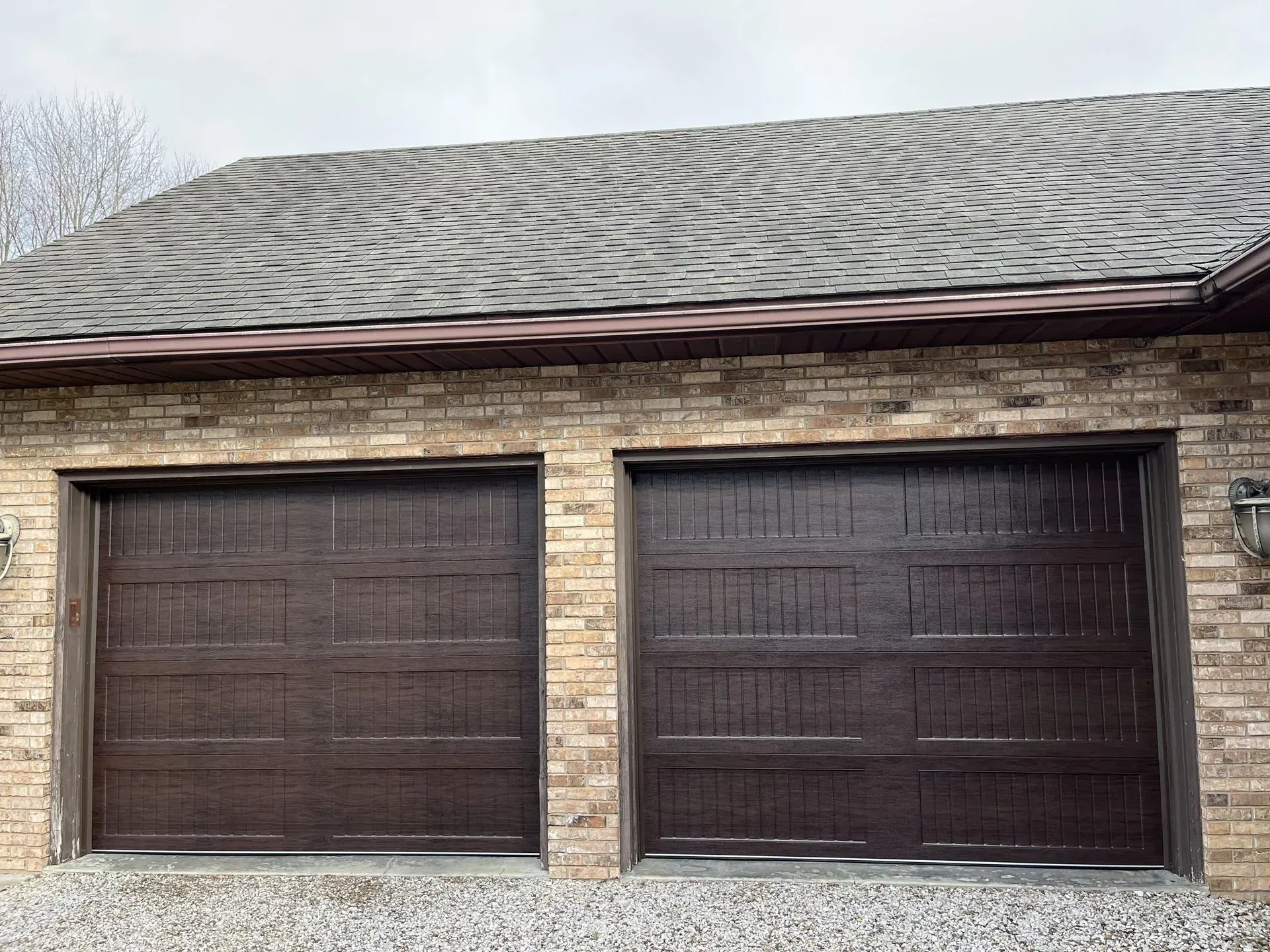 Two dark brown garage doors on a brick house under a shingled roof with swirling patterns