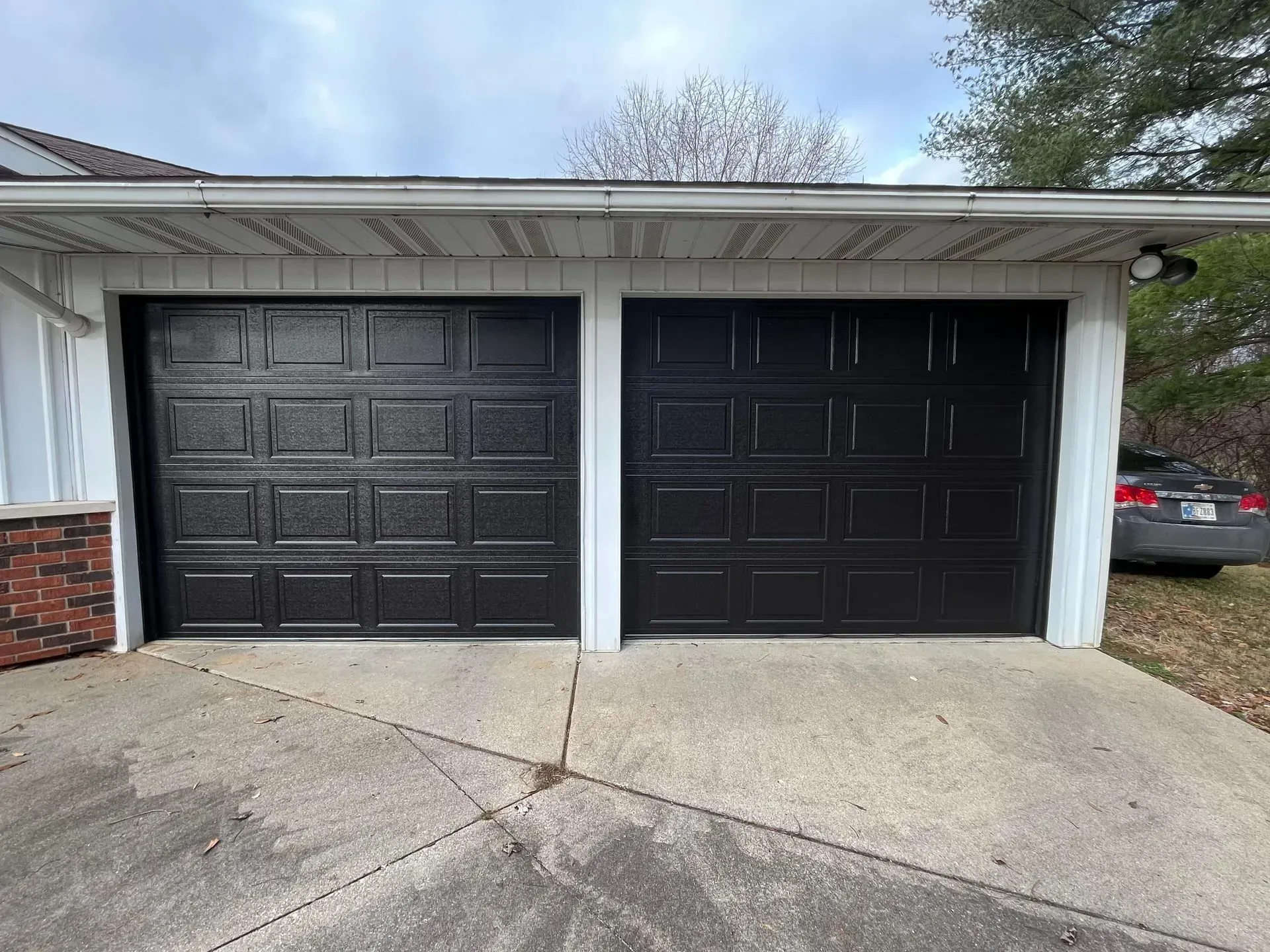 Two black garage doors in a white house exterior with a concrete driveway under a cloudy sky