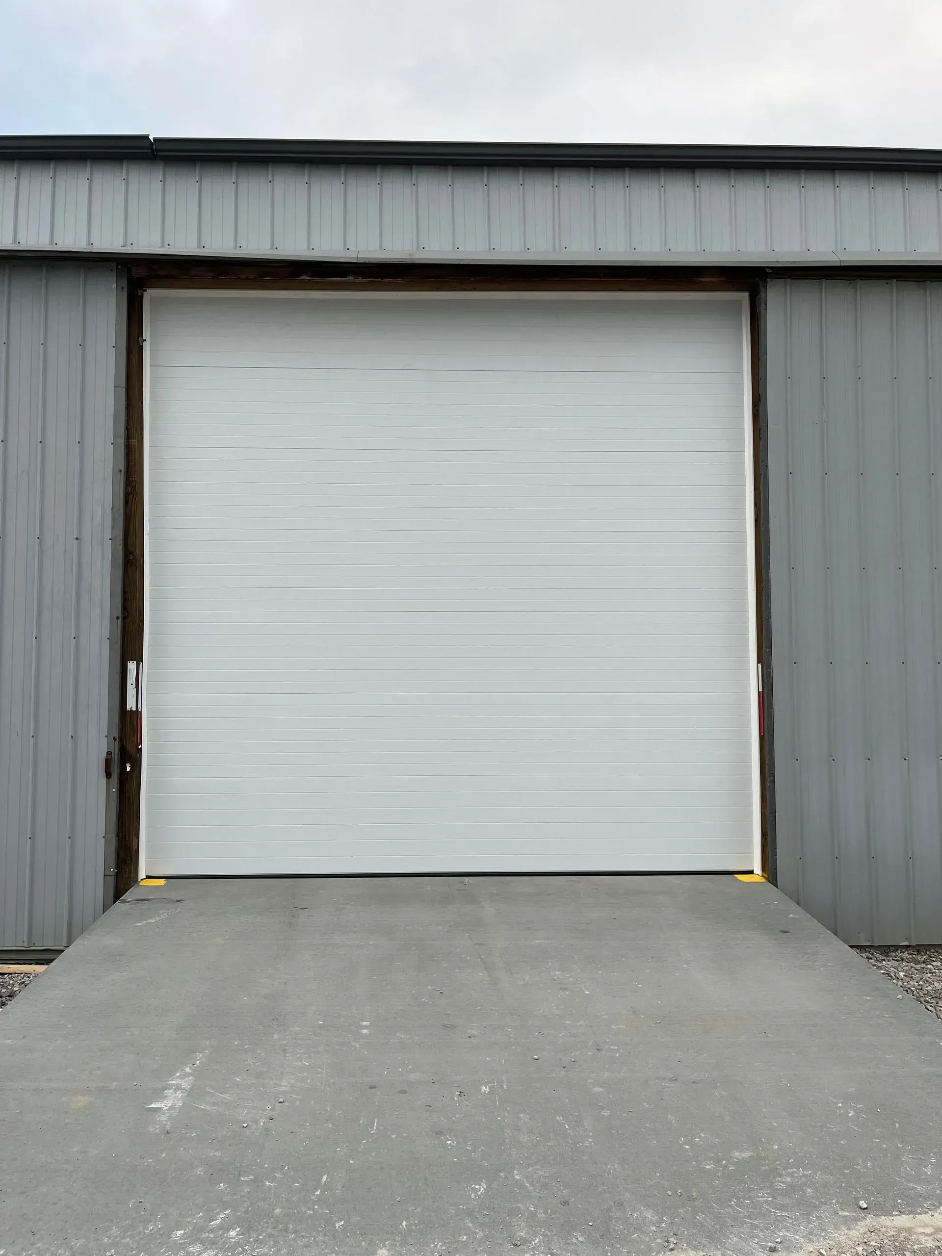 Closed white roll-up garage door on a gray metal building with a concrete driveway.