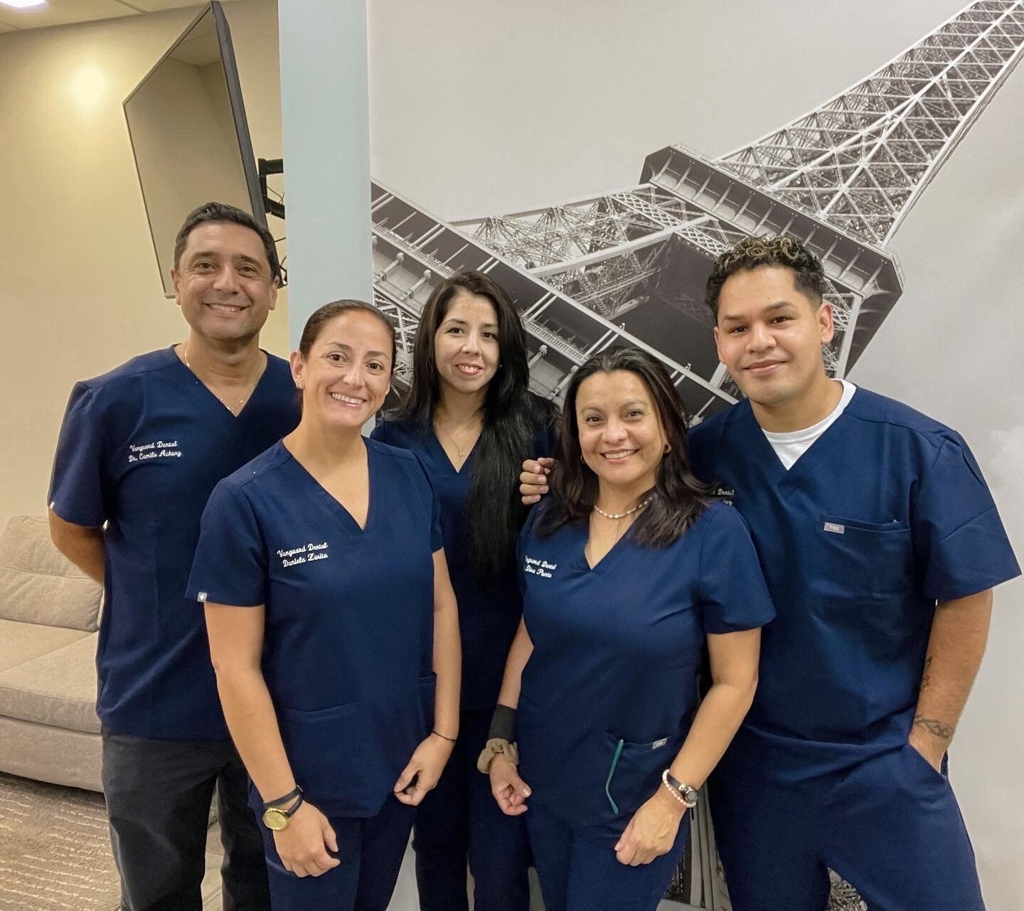 Group of five medical professionals in blue scrubs pose in front of Eiffel Tower graphic.