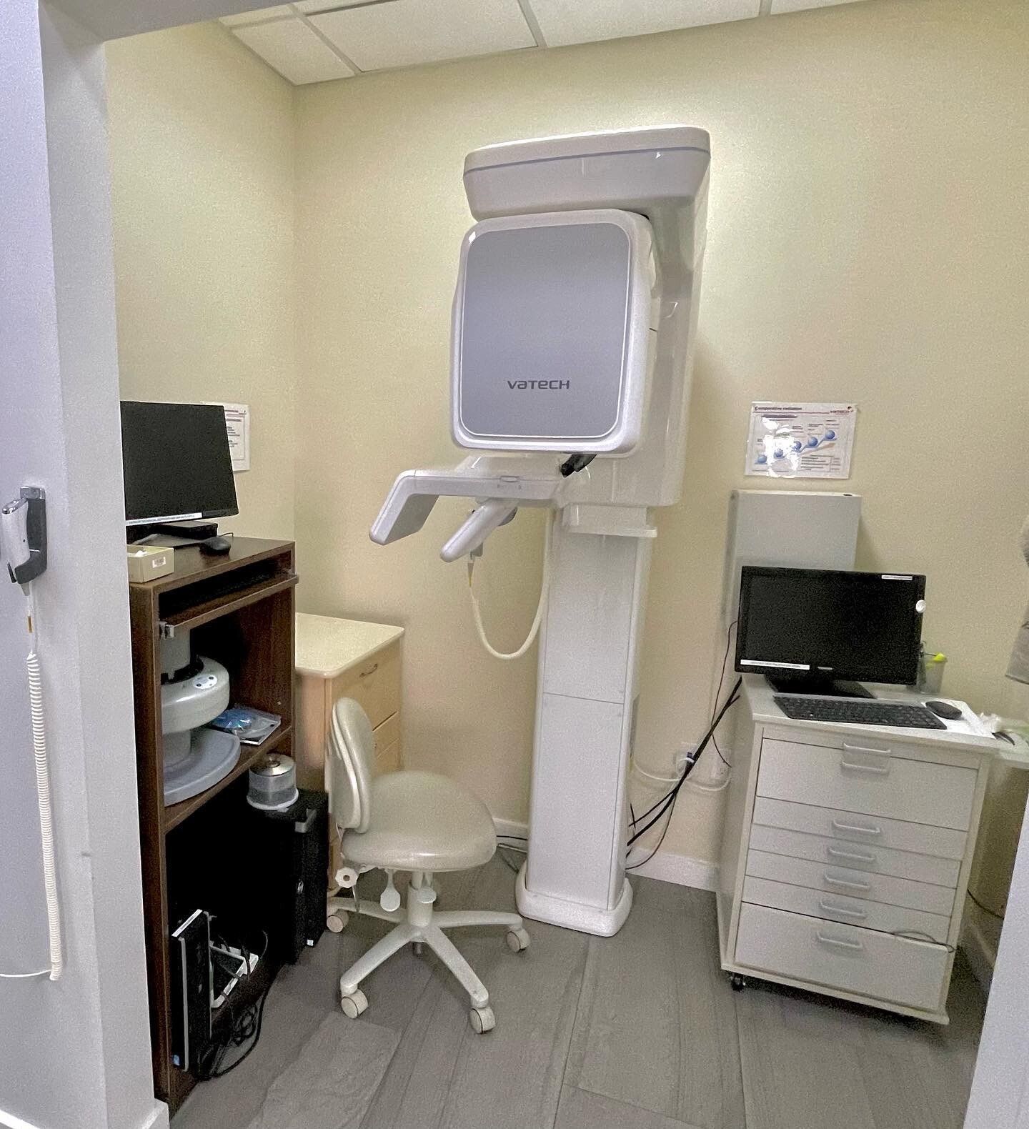 Dental X-ray room with scanning machine, chair, computer, and filing cabinet; walls are beige.
