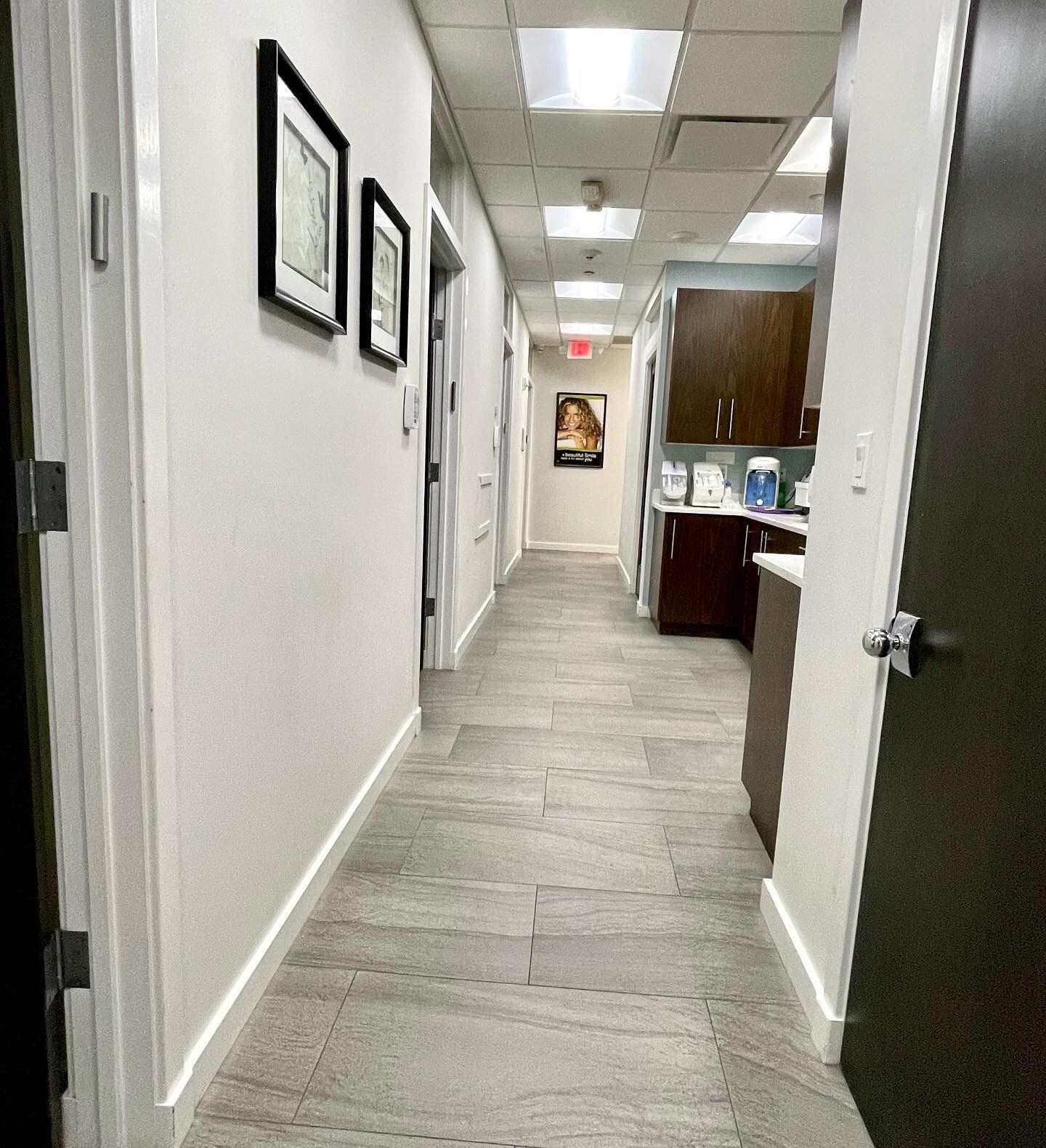 Narrow hallway in a medical office with gray tile floor, white walls, and framed artwork.