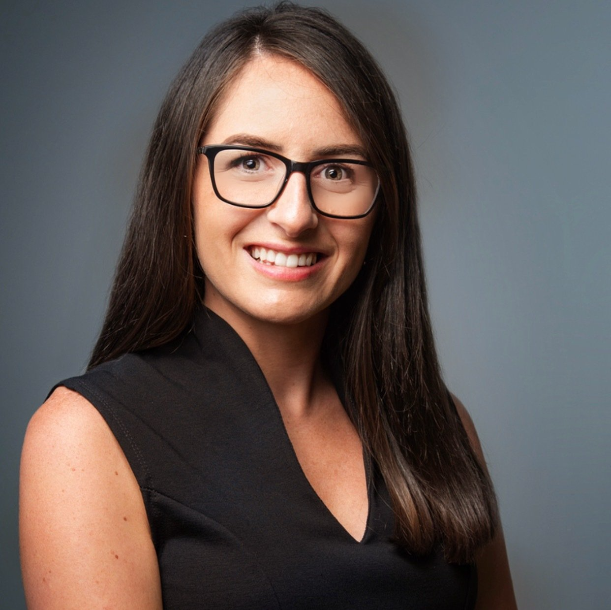 Smiling person with long dark hair and glasses, wearing a black sleeveless top against a gray background