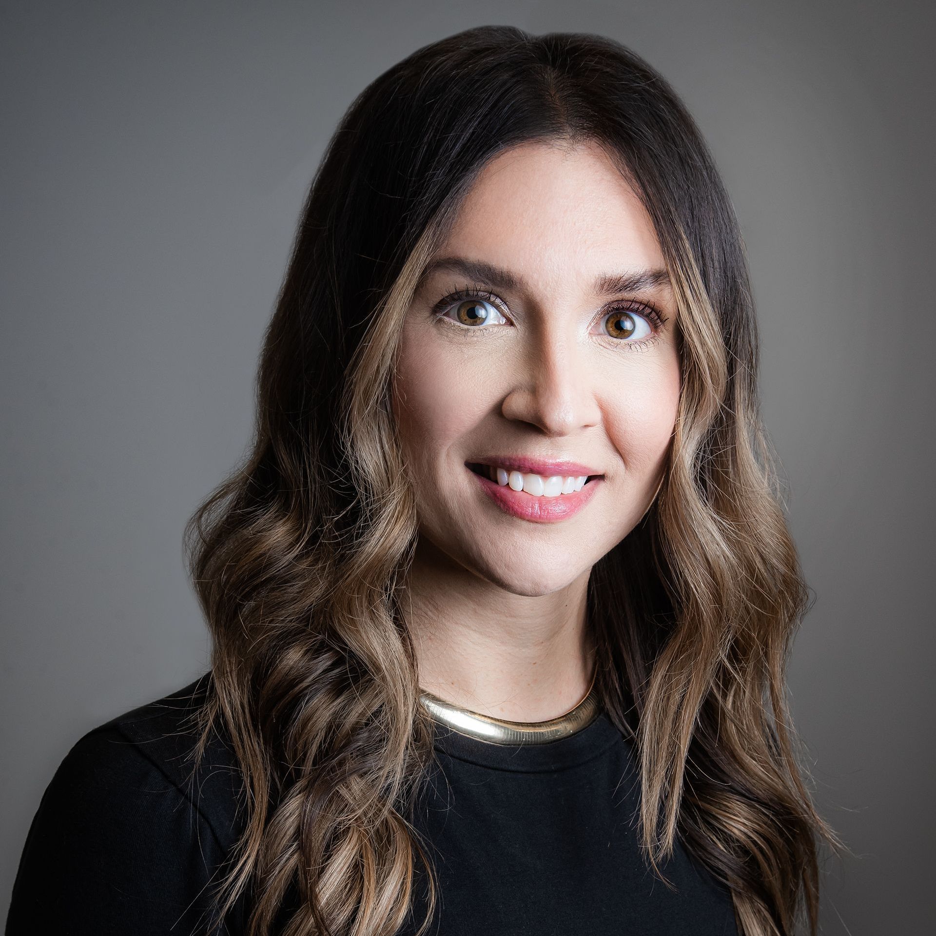 Studio portrait of a woman with long wavy hair, wearing a black top and smiling against a gray background