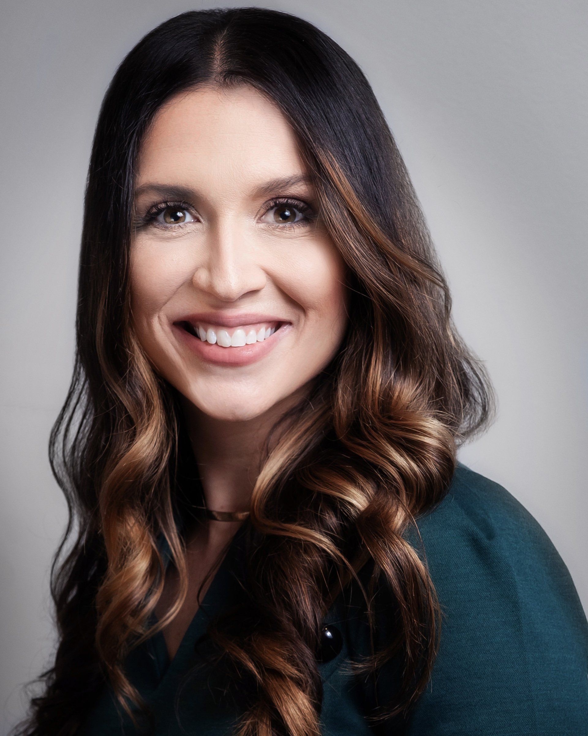 Smiling woman with long wavy brown hair on a gray background, wearing a dark top