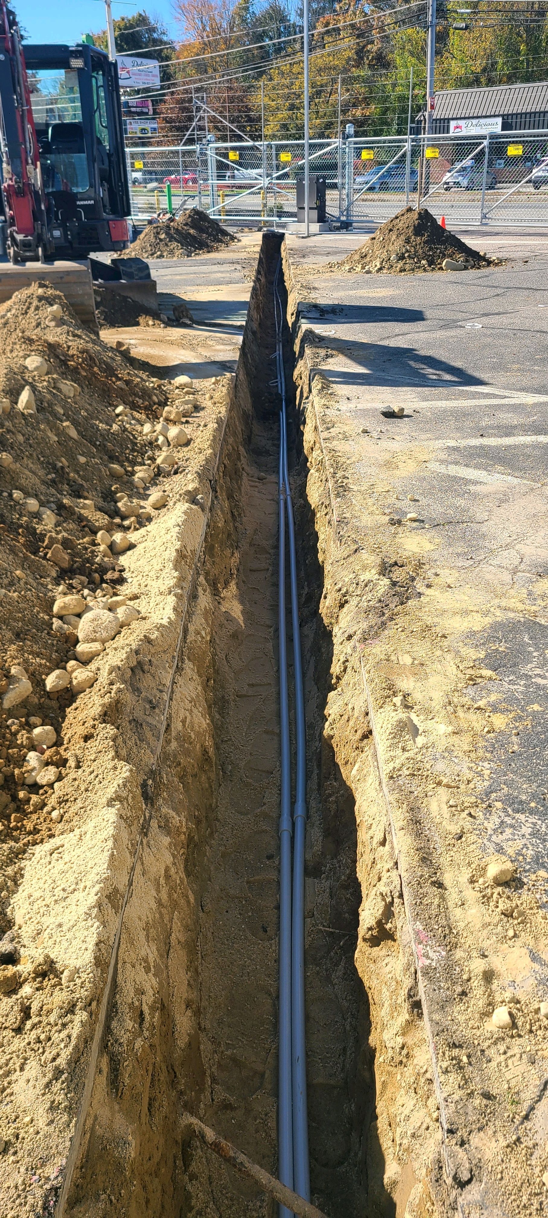 Trenched utility line beside a construction site, with excavator and gravel piles in the background.