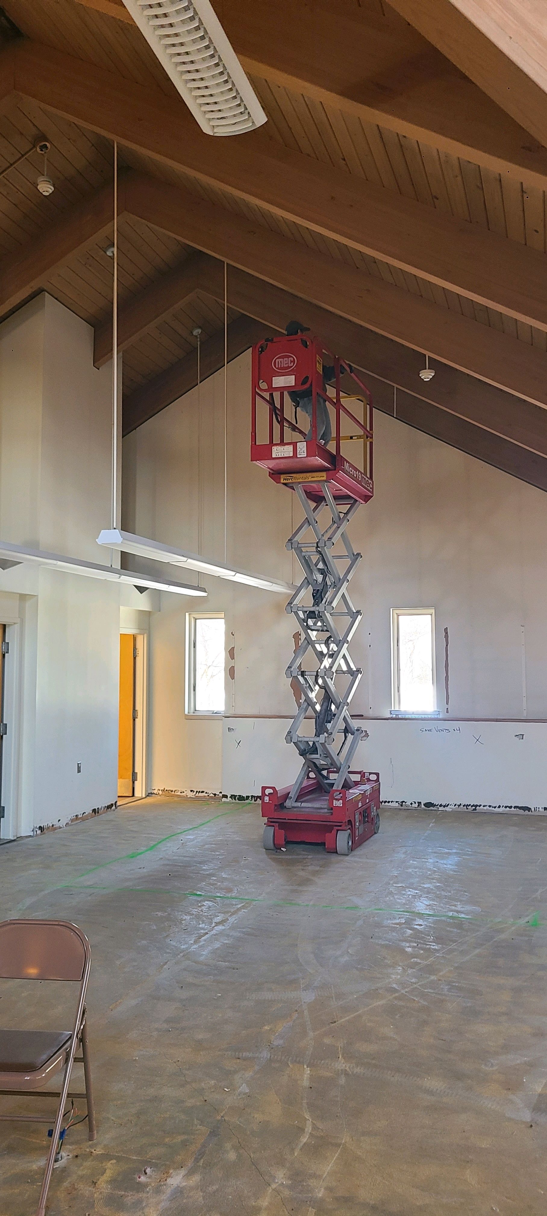 Red scissor lift extended inside an unfinished room with exposed wooden ceiling beams and concrete floor.