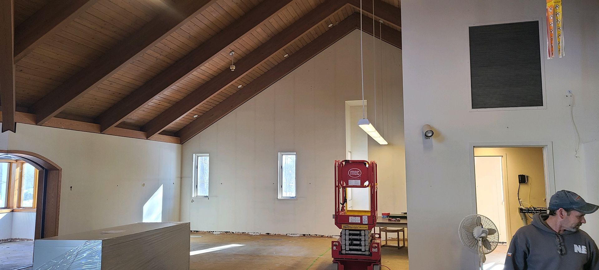Large room with high wooden ceiling, white walls, windows, and a red heater; a person stands at the doorway.