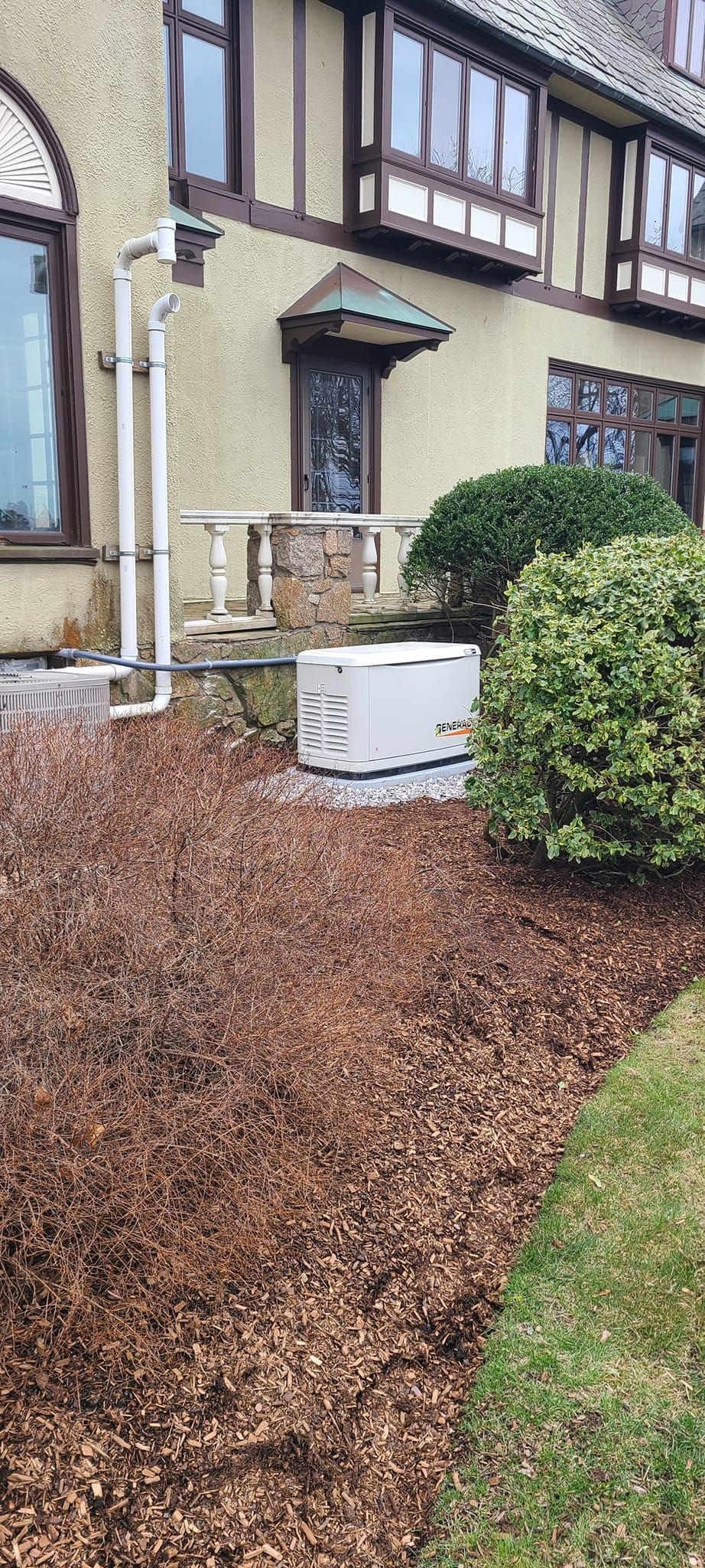 House side yard with white utility box, green shrubs, and brown mulch near a beige wall