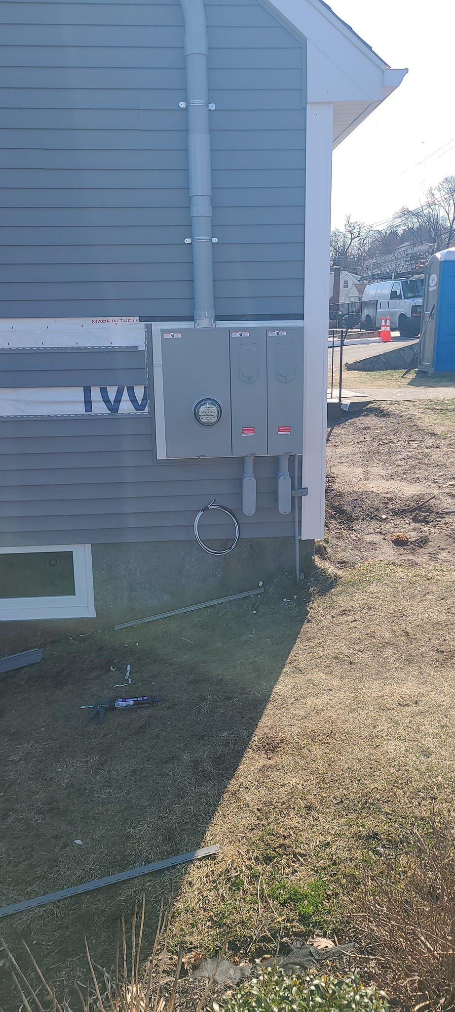 Gray utility boxes mounted on a house exterior beside a grassy yard and dirt driveway