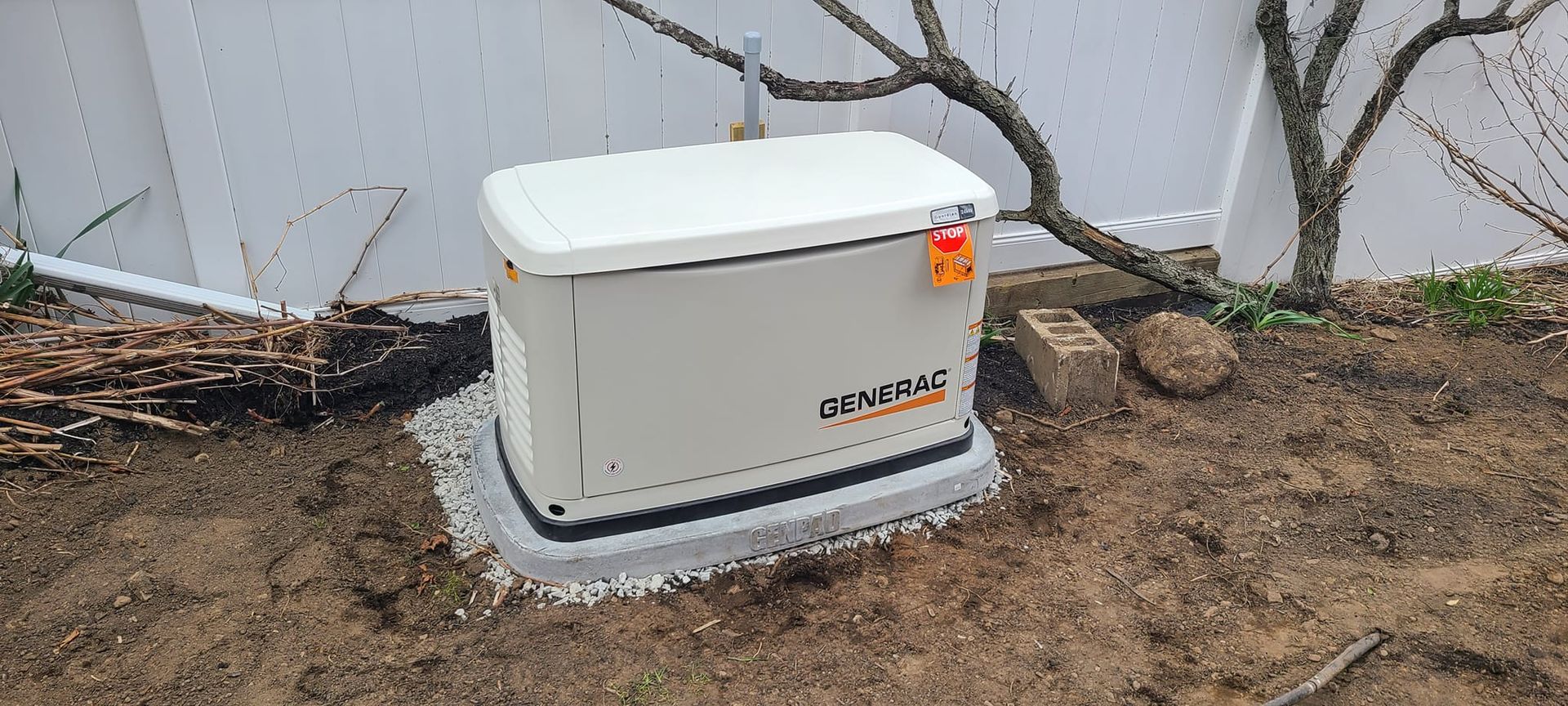 White utility generator on a concrete pad beside a fence and bare trees in a yard