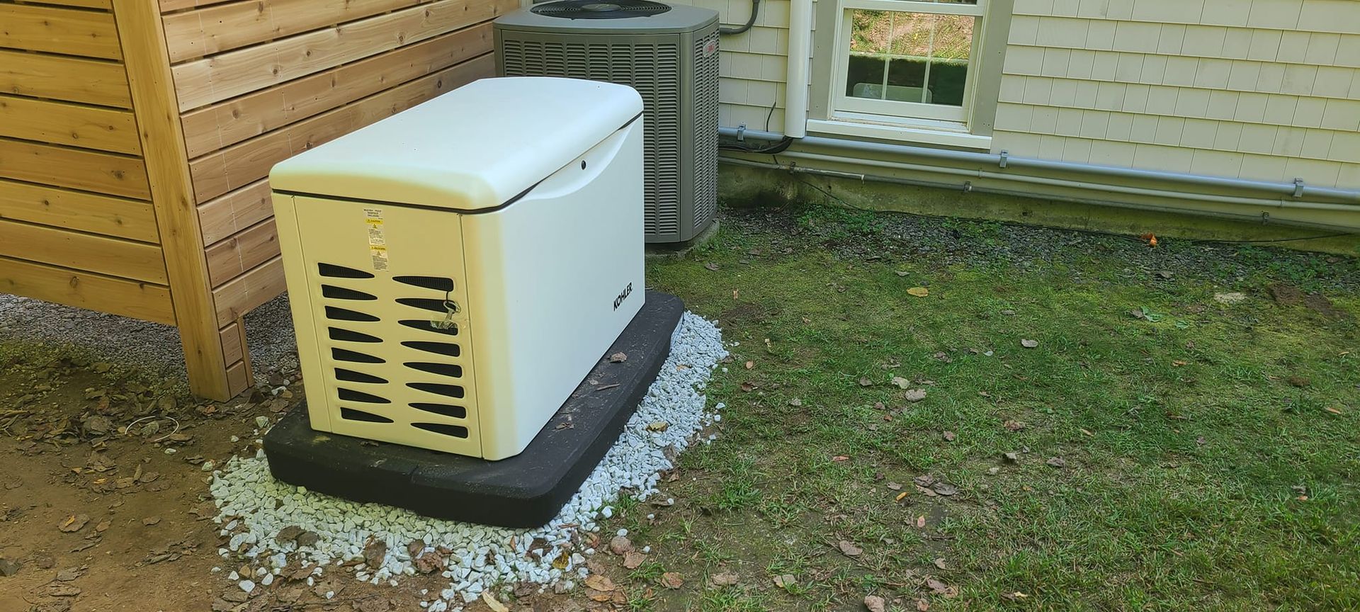 Outdoor generator beside a wooden building on a gravel base, with ventilation grilles and an extension cord behind it
