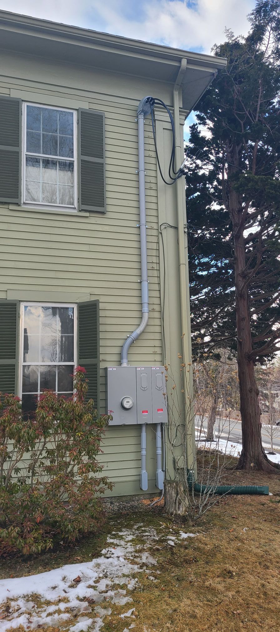 Side of a pale green house with white-trim windows, utility boxes, and snowy ground beside tall trees.