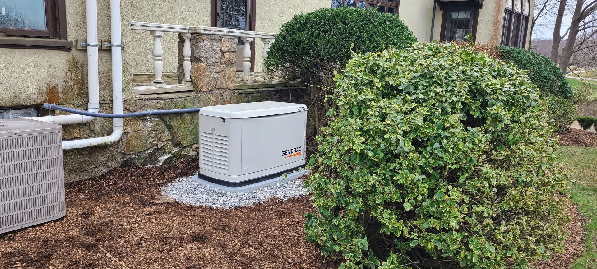 White outdoor HVAC unit beside a building, partly hidden by green shrubs and mulch.