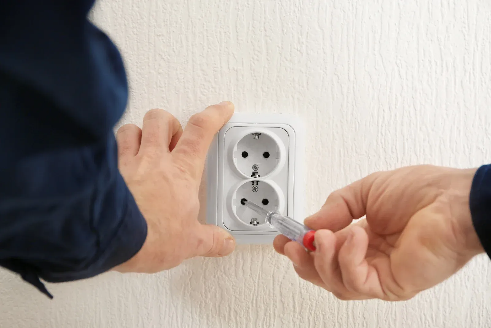 Hands using a screwdriver to install a white double wall outlet on a textured wall