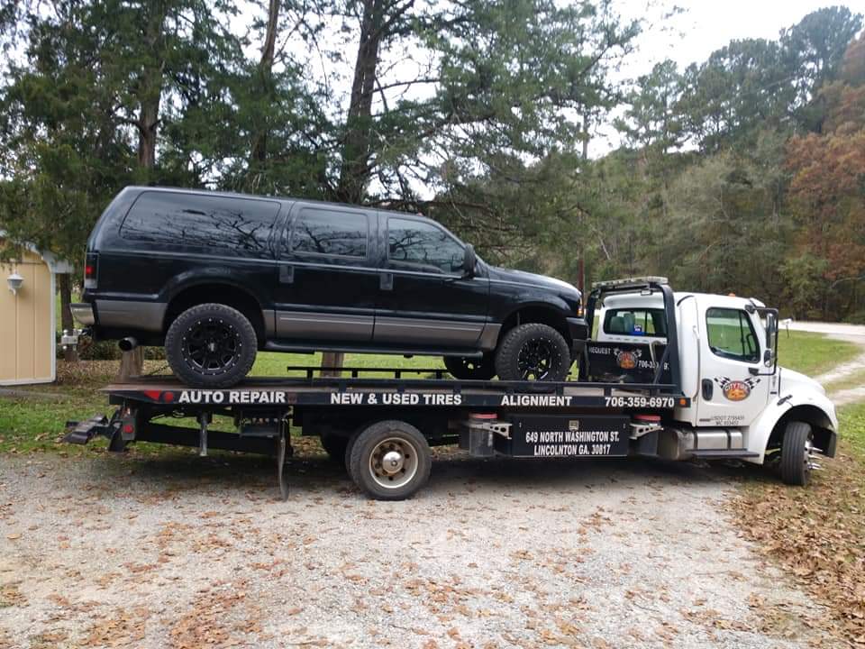Black SUV on tow truck; parked in front of a small building and trees.