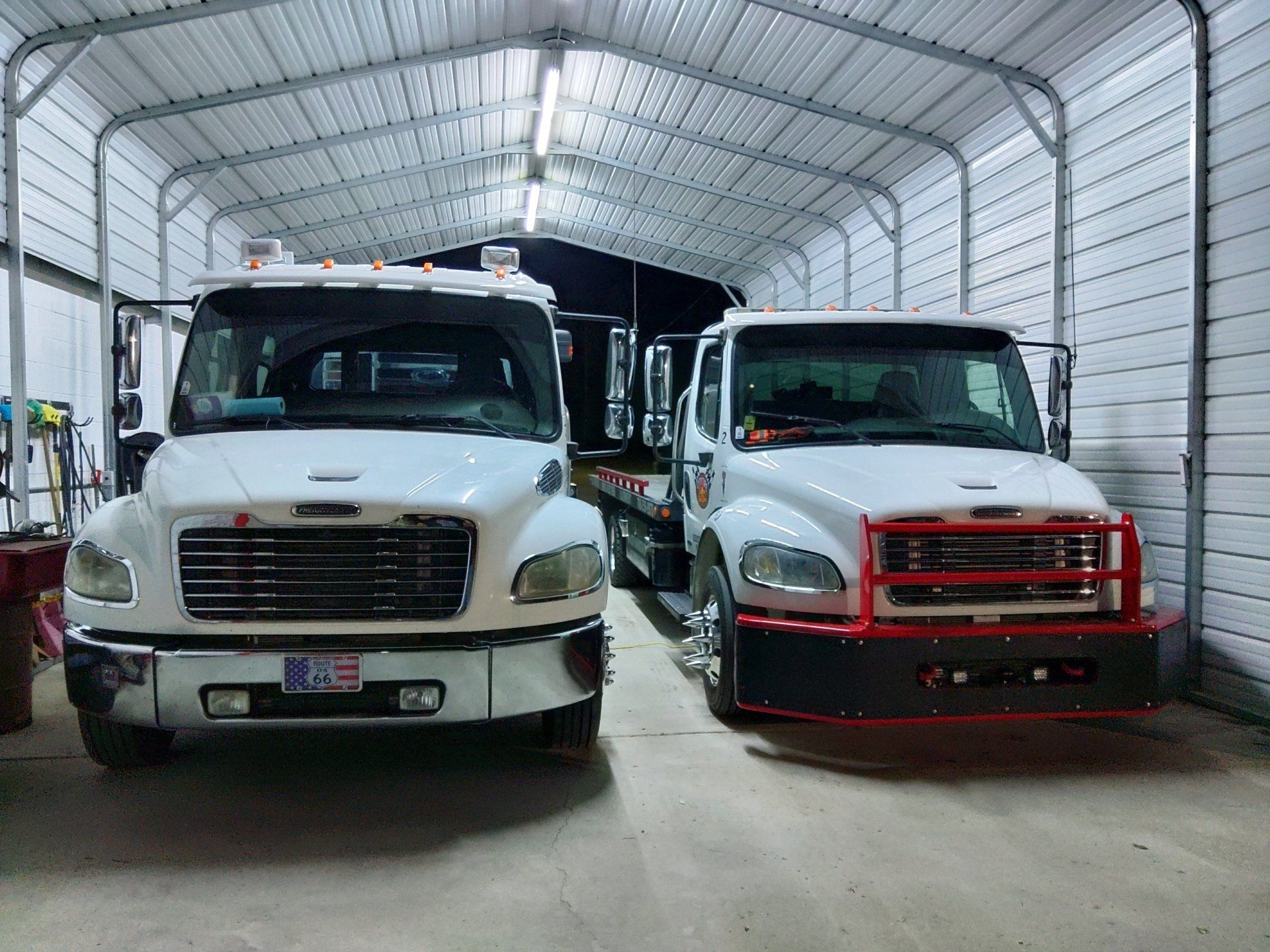 Two white tow trucks inside a metal garage. One has a chrome grill, the other a red bumper guard.