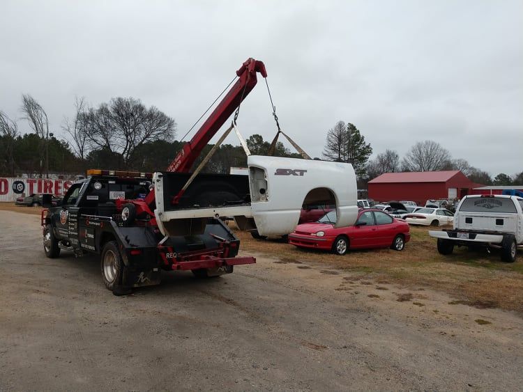 Tow truck lifting a white truck bed in a yard, red car nearby, overcast day.