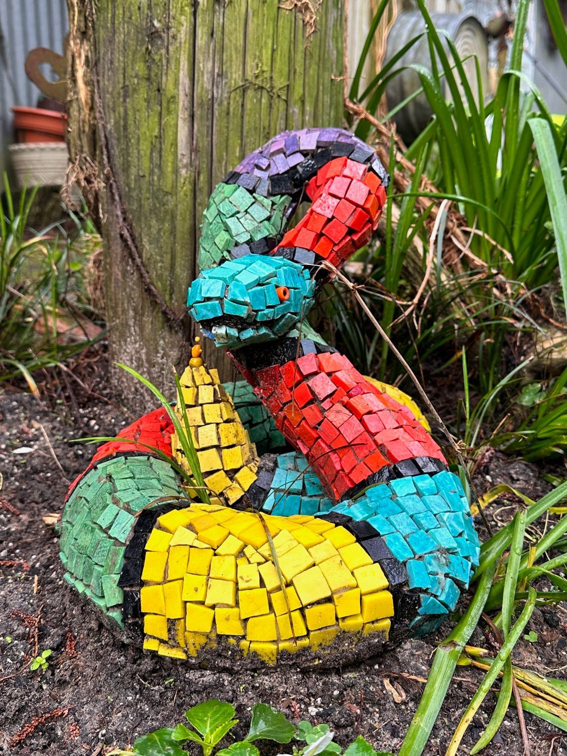 A colorful mosaic snake is sitting in the dirt next to a tree.