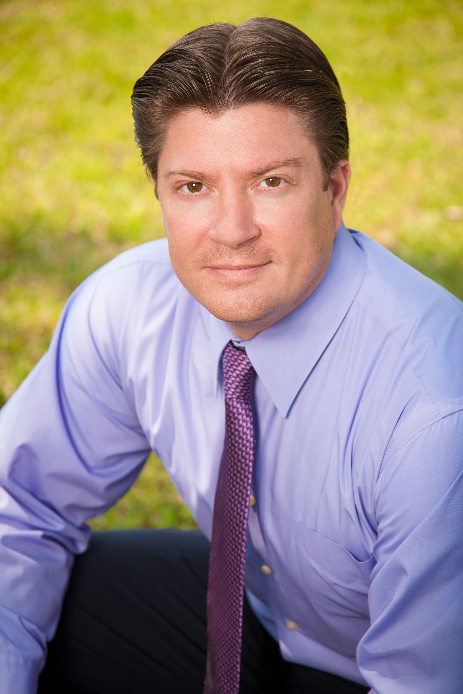 Man in a blue shirt and purple tie, smiling, sitting outdoors in front of green grass.