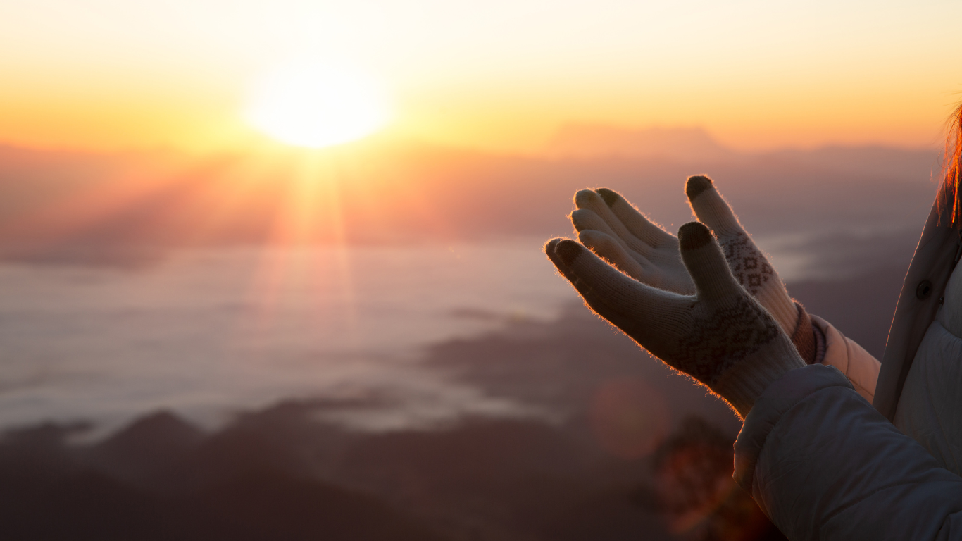 Hands outstretched towards a bright sunrise, silhouetted on a mountain with a cloudy horizon.
