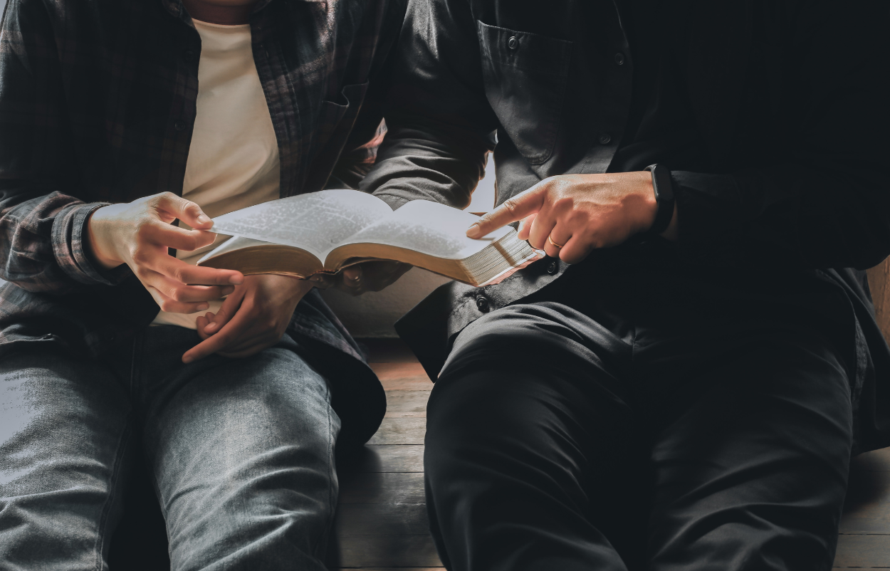 Two people sitting closely, reading an open book together. One points to text with a finger.