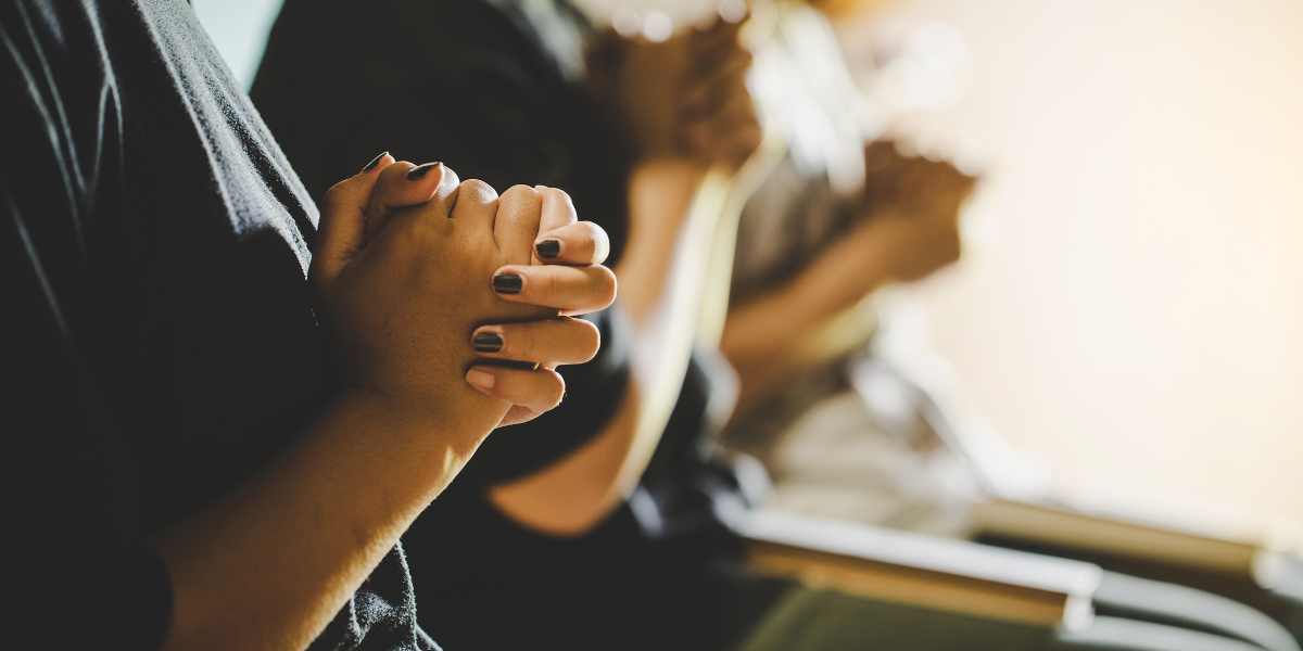 Hands clasped in prayer, with blurred figures in the background.