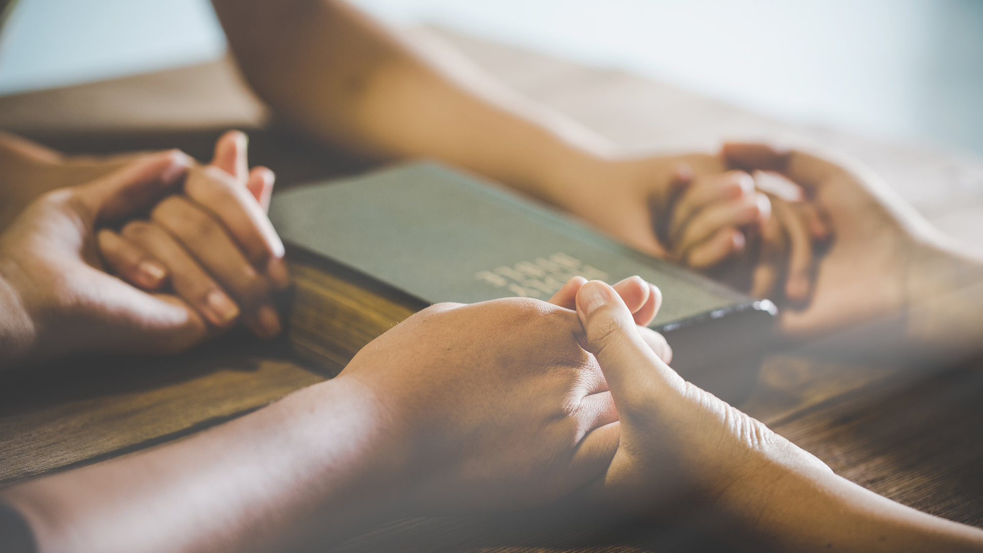 Hands clasped in prayer around a closed Bible on a wooden surface.