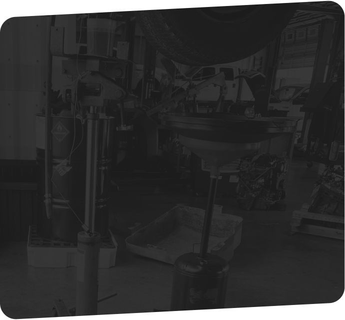 An oil drain pan on a stand positioned beneath a lifted vehicle in a dark, cluttered garage setting.