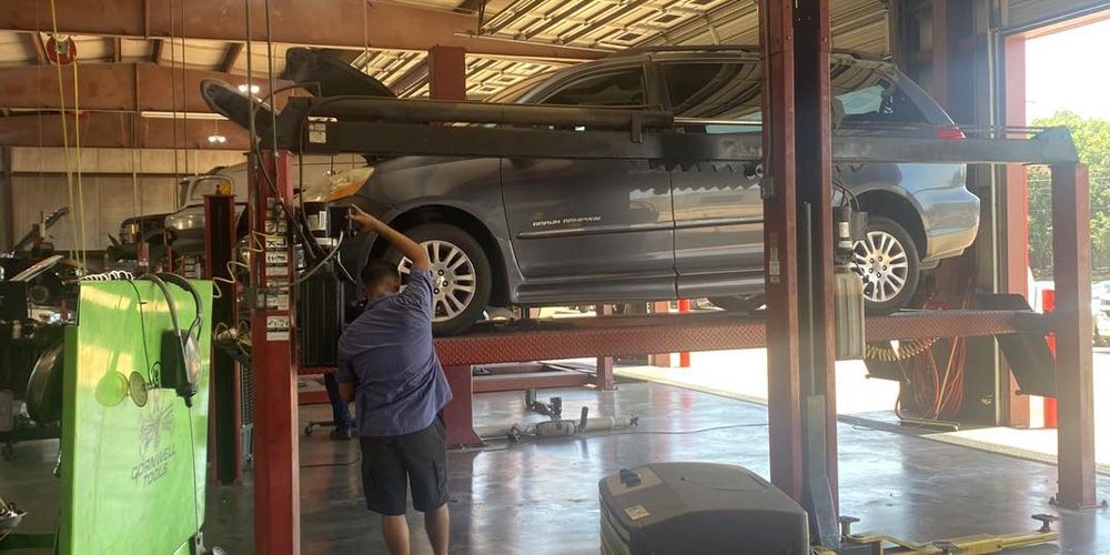 A person in a blue shirt works on a dark car raised on a hydraulic lift inside an auto repair shop.