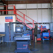A wide shot of an automotive shop interior with a diagnostic machine, red stairs leading to a mezzanine, and tool carts.