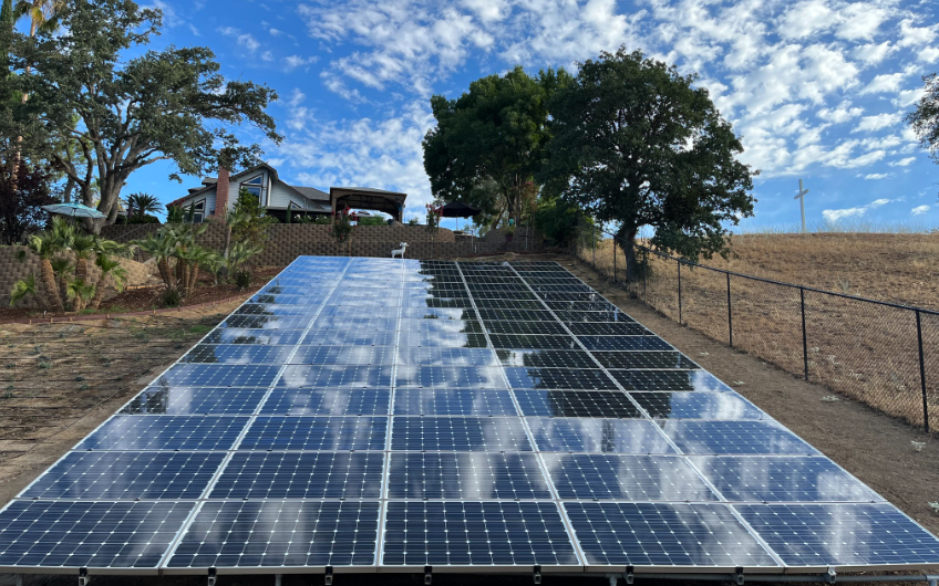 A row of solar panels are sitting on top of a dirt field in front of a house.