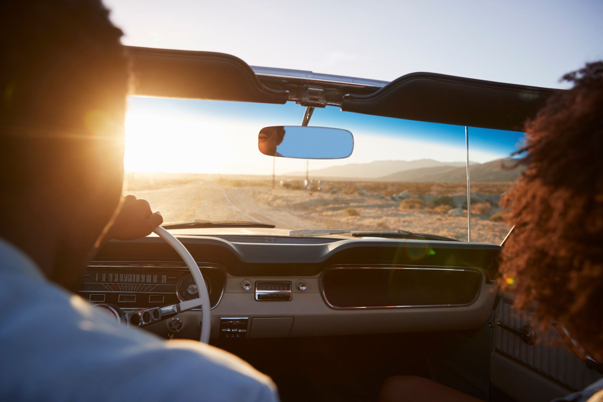 Two people in a convertible driving into a bright sunset over a desert road