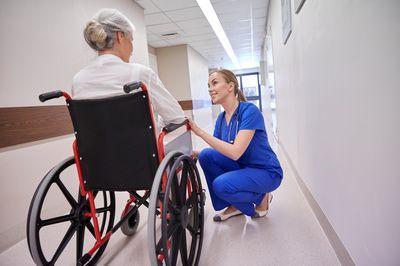 A nurse is kneeling down next to an elderly woman in a wheelchair in a hospital hallway.